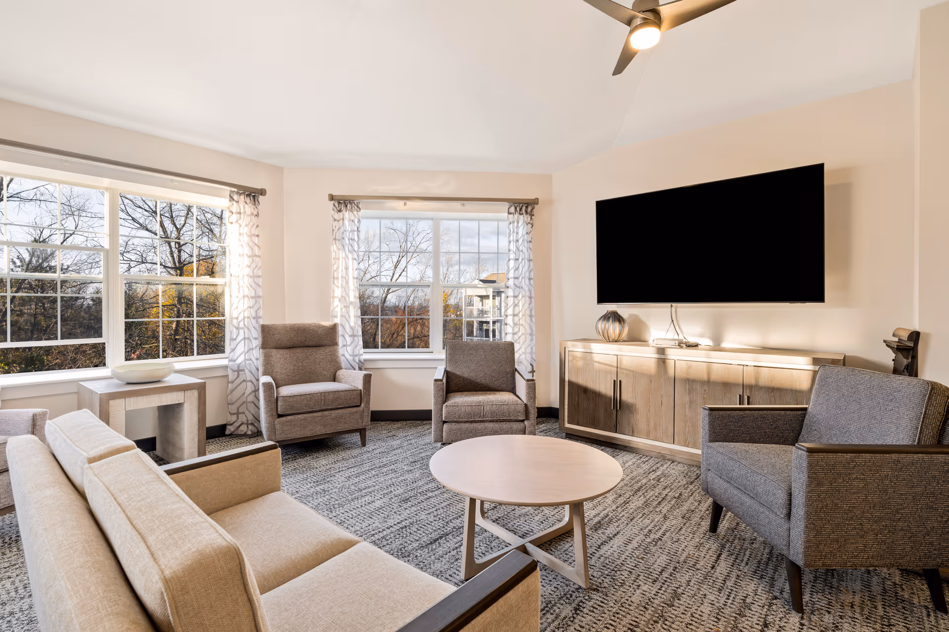 A bright and modern living room in a senior living community featuring a beige sofa, three gray armchairs, a round wooden coffee table, a wooden cabinet with a large flat-screen TV mounted above it, and large windows with patterned curtains letting in natural light.