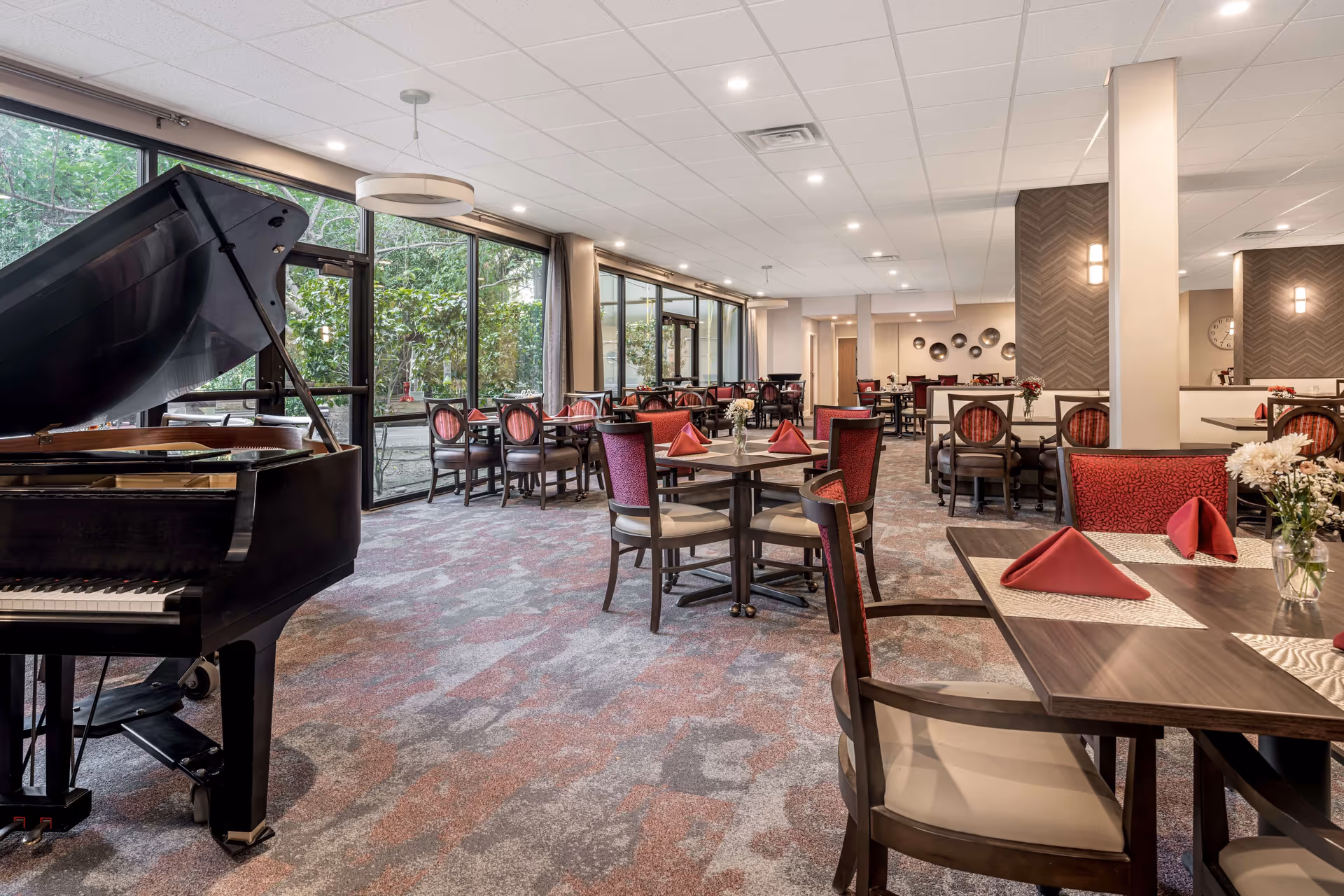 A spacious dining room with multiple tables set with red napkins and floral centerpieces. There is a black grand piano on the left side near large windows that show greenery outside. The room has carpeted floors, modern lighting, and a warm, inviting atmosphere.
