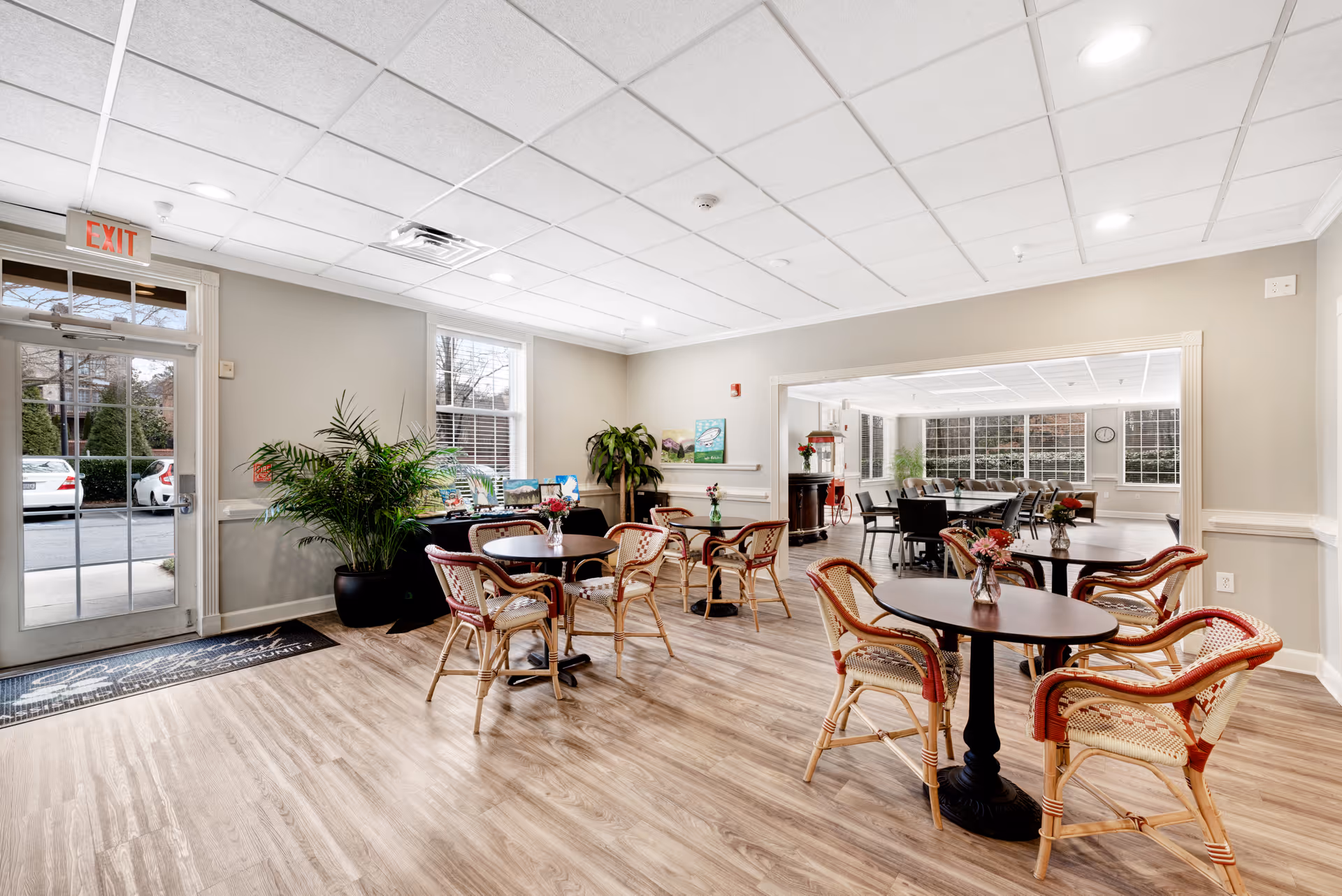 A bright and spacious common area in a senior living facility with several round tables and wicker chairs arranged for seating. There are plants near the windows and a glass door leading outside. The room has light-colored walls, wood flooring, and a drop ceiling with recessed lighting. In the background, there is an adjoining room with more tables and chairs, and a popcorn machine is visible.