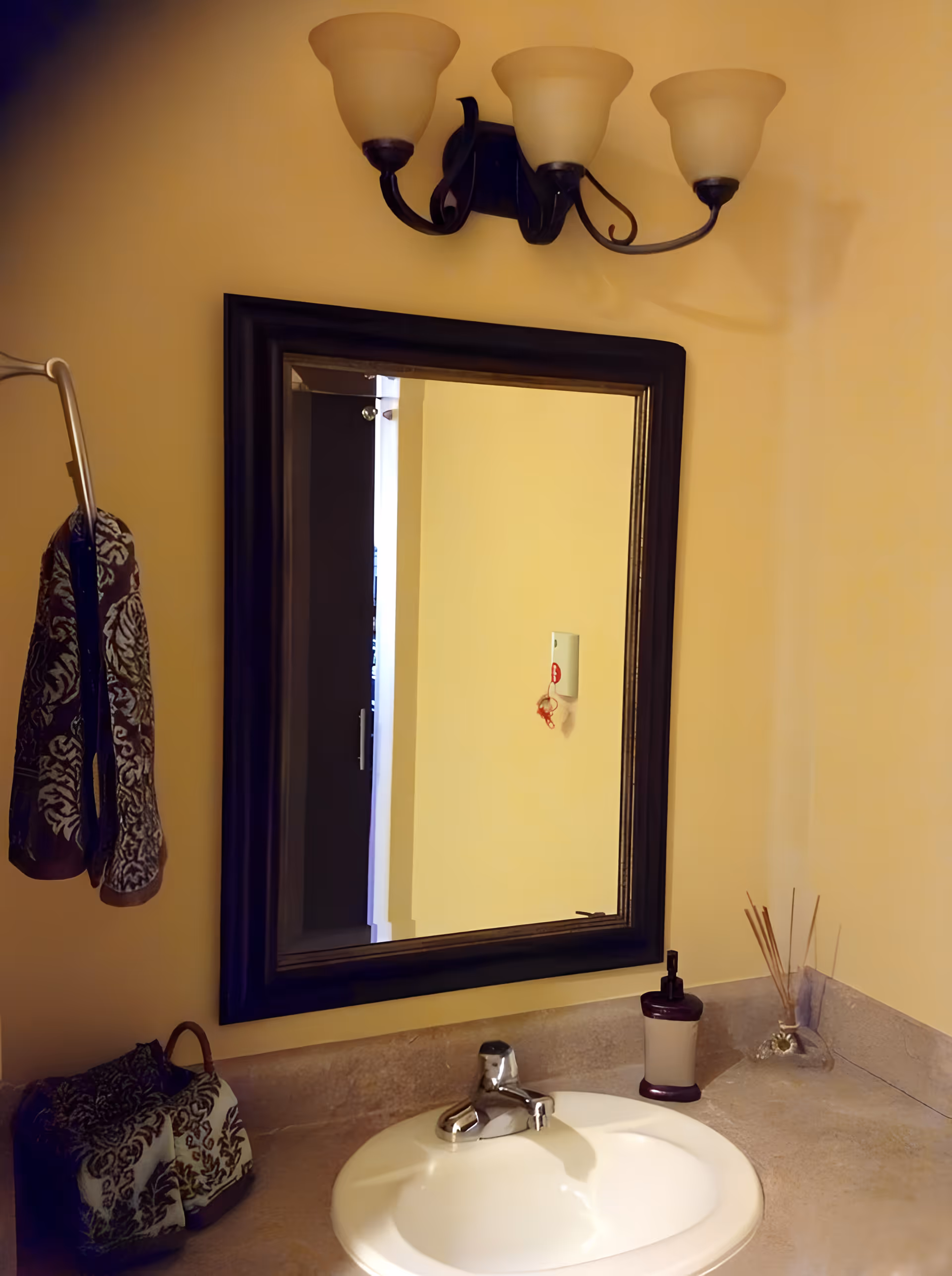 A bathroom sink area with a white oval basin and a silver faucet set in a beige countertop. Above the sink is a large rectangular mirror with a dark frame, reflecting a yellow wall and a door. Above the mirror is a three-light wall sconce with frosted glass shades. To the left, a towel rack holds two patterned towels, and on the countertop are a matching soap dispenser and a reed diffuser.
