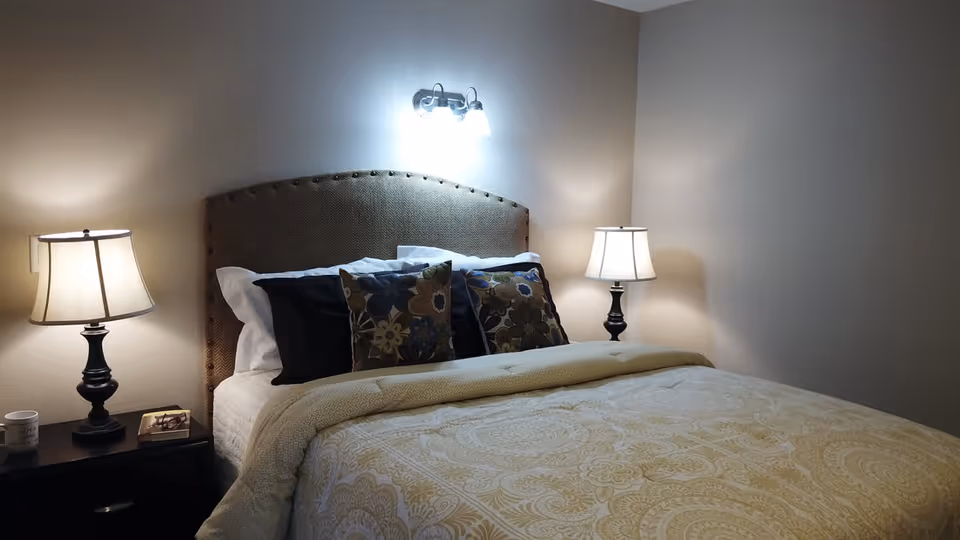 A neatly made bed with a patterned beige comforter and multiple pillows, including floral decorative pillows, against a beige wall. There are two bedside tables, each with a lamp, and a wall-mounted light fixture above the headboard.