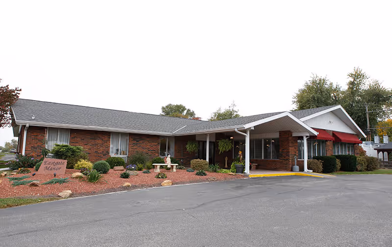 Exterior view of Eastgate Manor Nursing & Rehabilitation, a single-story brick building with a gray shingled roof, a covered entrance supported by white columns, red awnings over windows, and landscaped grounds with bushes and a stone sign displaying the facility's name.