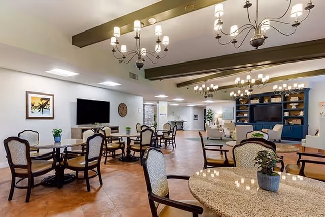 Open communal dining and lounge area with round tables and chairs, chandeliers, and a television wall in a senior living facility.
