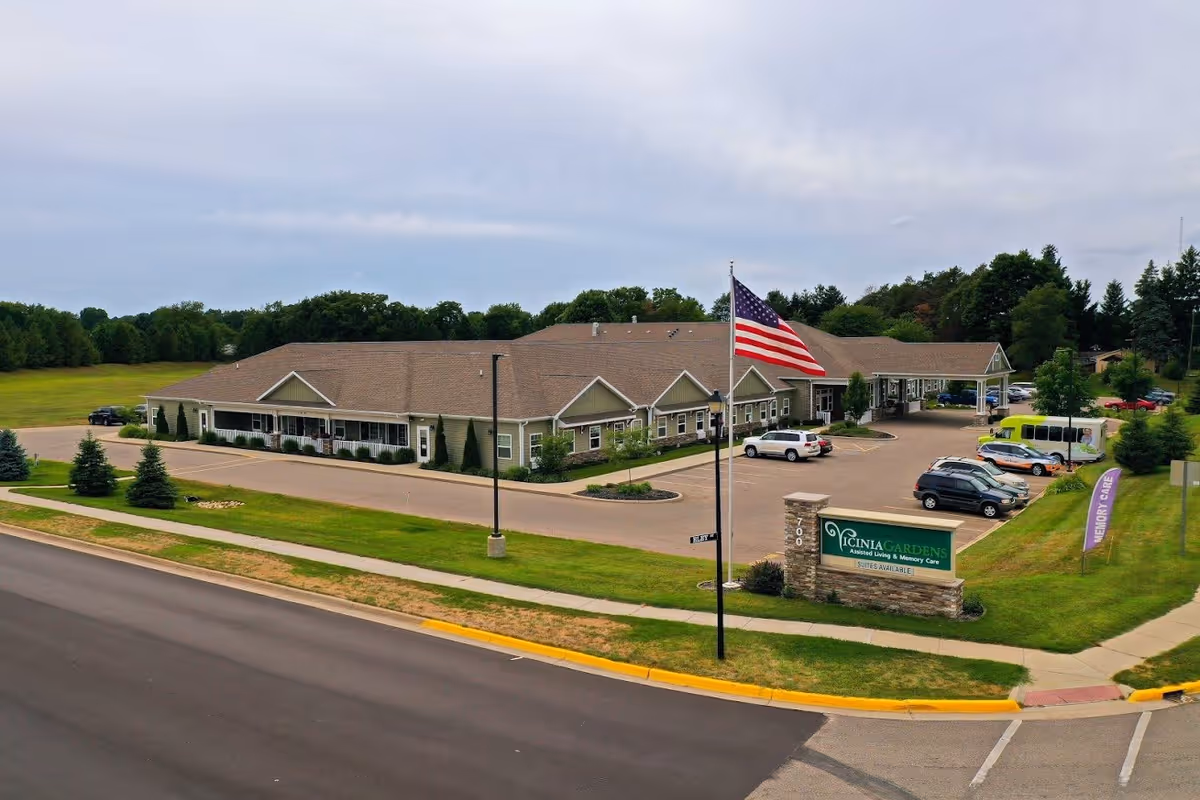 Exterior view of a single-story assisted living and memory care facility with a large parking lot, an American flag on a flagpole, and a green sign that reads 'Vicinia Gardens Assisted Living & Memory Care Suites Available'. The building is surrounded by grass and trees under a cloudy sky.