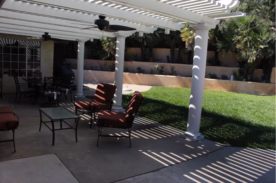 Outdoor patio area with white pergola supported by columns casting striped shadows on the ground. There are cushioned chairs and a glass-top table under the pergola, with a grassy lawn and plants in the background.