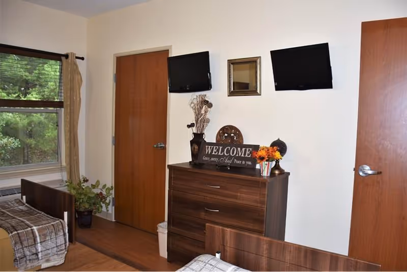 A cozy room with two wooden beds covered in plaid blankets, a wooden dresser with decorative items including a welcome sign, a vase with dried flowers, and a small flower arrangement. Two flat-screen TVs are mounted on the wall above the dresser. There are two wooden doors and a window with beige curtains showing greenery outside.