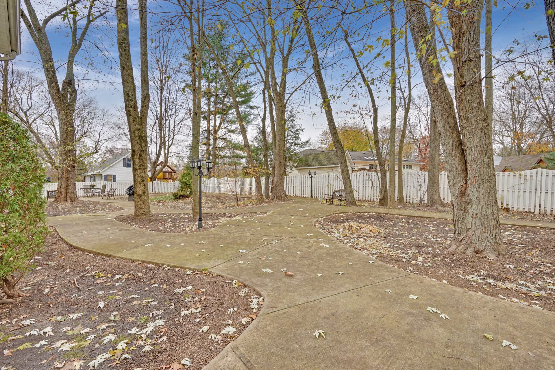Outdoor paved walking paths surrounded by leafless trees and a white picket fence, with benches and a lamp post along the paths under a partly cloudy sky.