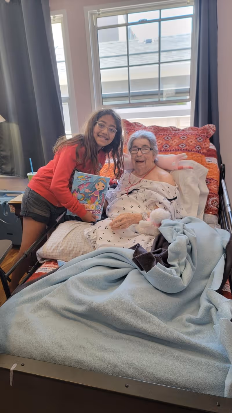 An elderly woman lying in a hospital-style bed covered with a light blue blanket and a colorful pillow behind her. She is smiling and wearing glasses and a hospital gown. Next to her is a young girl with long hair and glasses, wearing a red sweatshirt and black shorts, holding a colorful book. They are in a room with two windows and dark curtains.