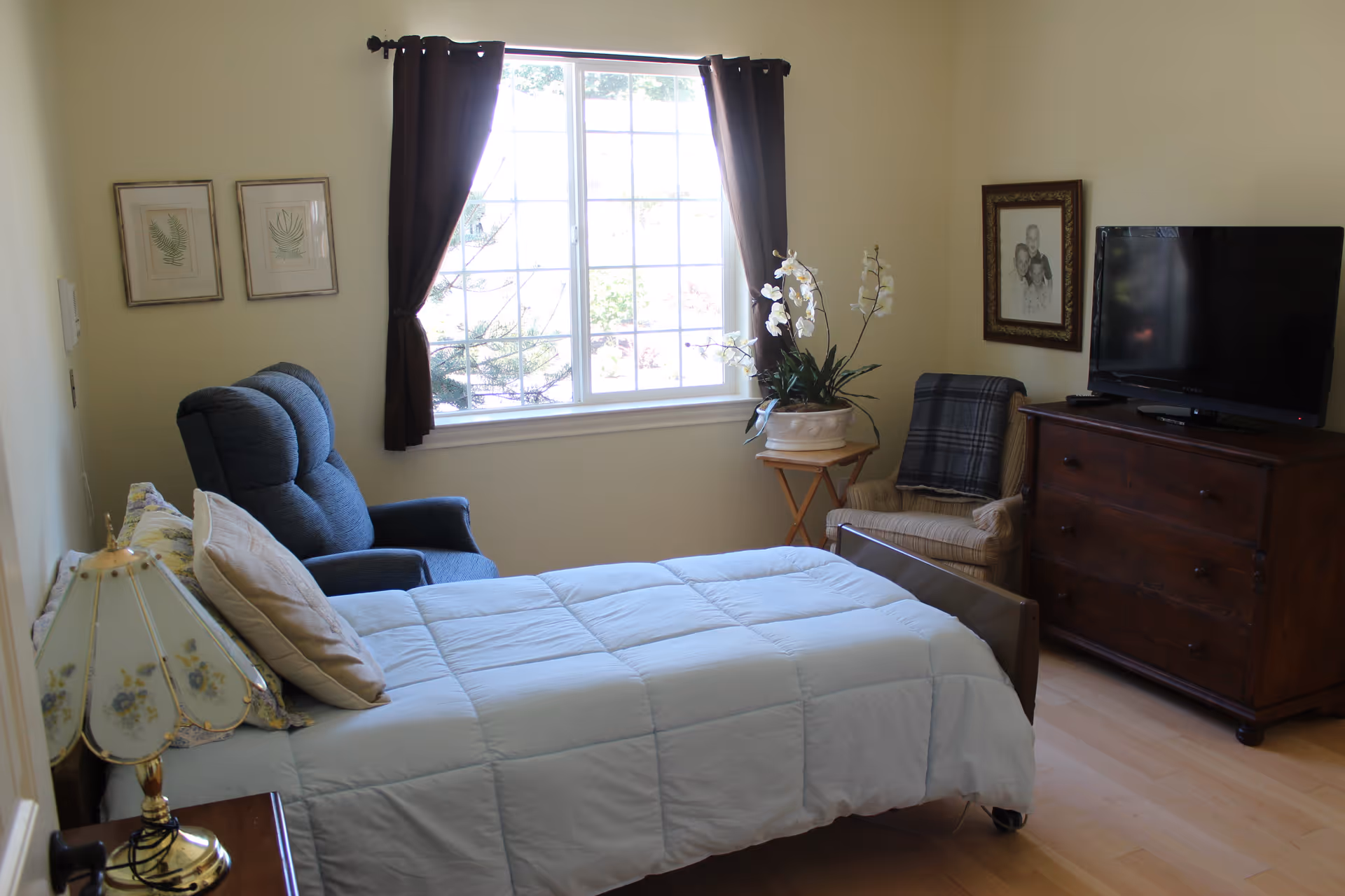A cozy bedroom with a single bed covered in a light blue comforter, a blue recliner chair, a wooden dresser with a flat-screen TV on top, a beige armchair with a plaid blanket, a small wooden side table with a white orchid plant, and a window with dark curtains letting in natural light. The walls are light yellow with framed artwork.
