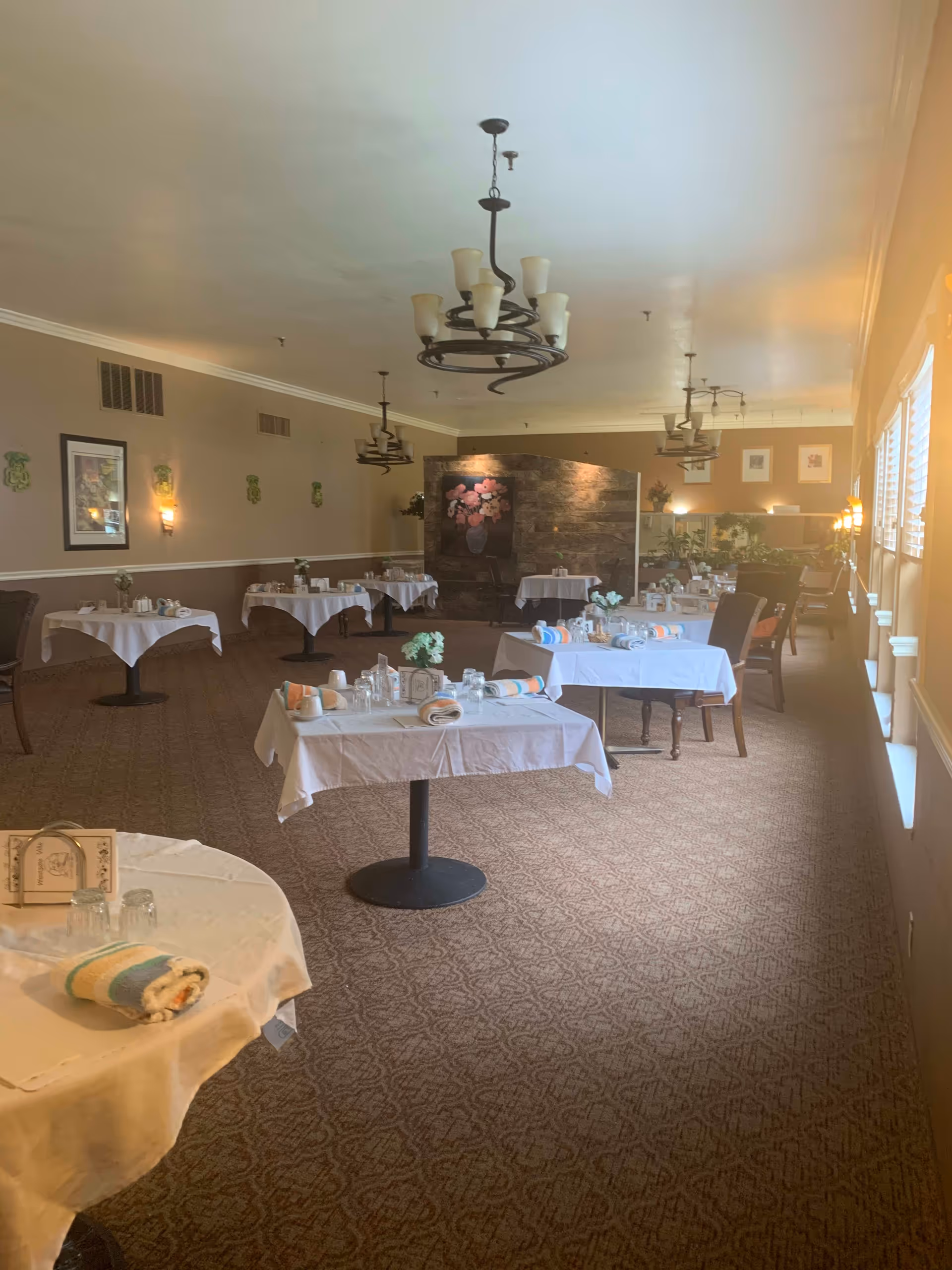 A dining room with multiple tables covered in white tablecloths, each set with rolled napkins, glasses, and small flower arrangements. The room has patterned carpet, beige walls with framed artwork, and several chandeliers hanging from the ceiling. Large windows on the right side allow natural light to enter the space.