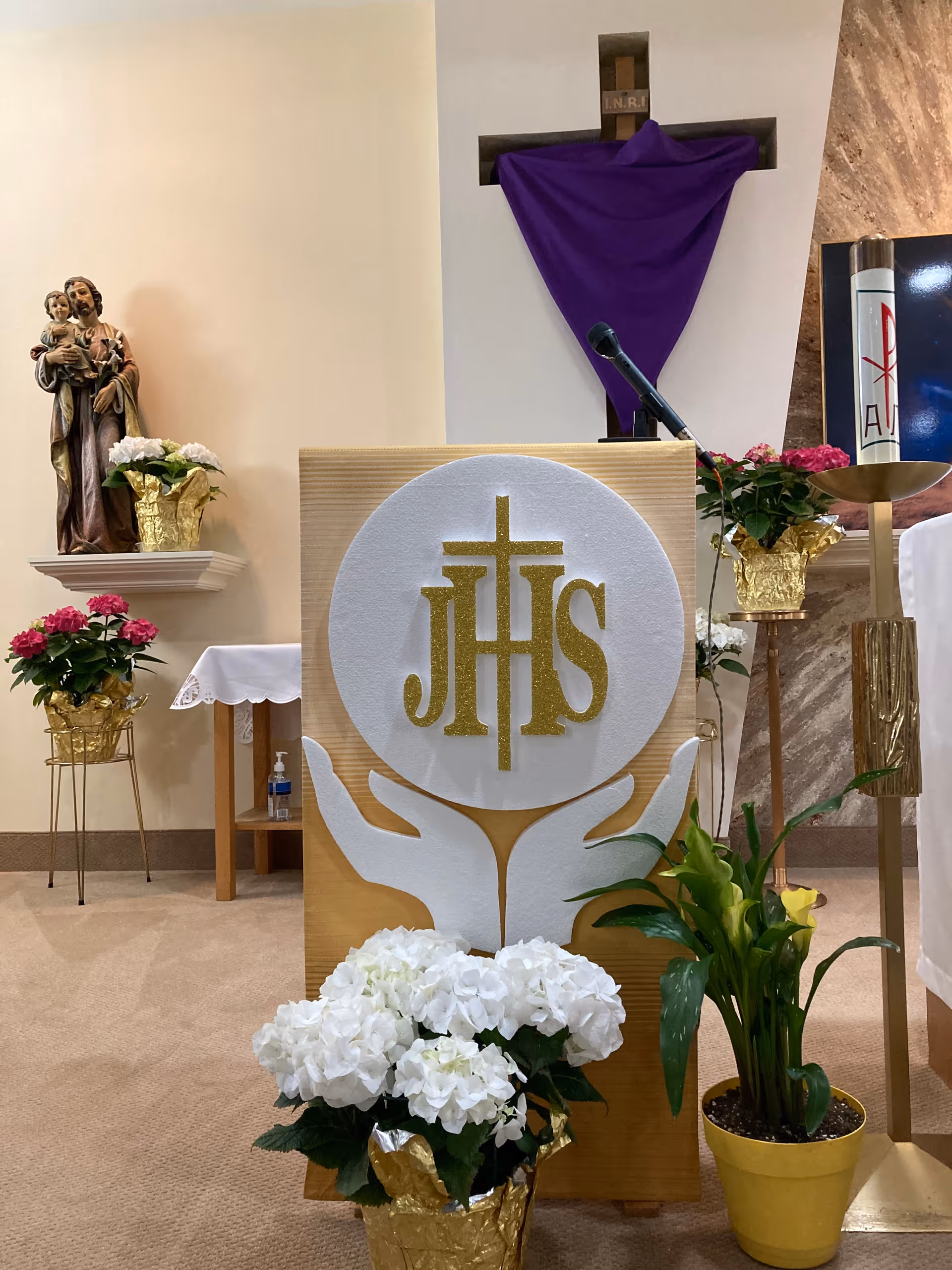 Interior view of a chapel area in a senior living facility featuring a wooden podium with a white and gold religious symbol 'JHS' and two white hands. Surrounding the podium are various potted flowers including white hydrangeas and yellow calla lilies. In the background, there is a statue of a saint holding a child on a wall shelf, a cross draped with a purple cloth, a microphone, and a tall candle with religious symbols.