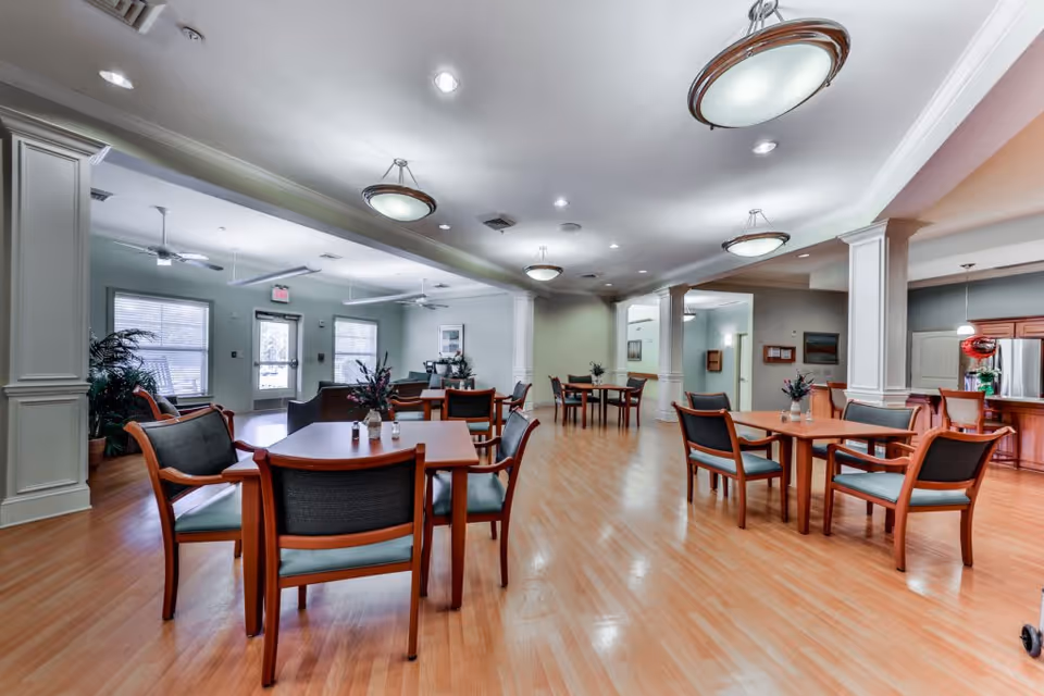 A spacious and well-lit common area in a senior living facility with wooden floors, multiple tables and chairs arranged for dining or socializing, ceiling fans, and decorative plants. The room has large windows allowing natural light and a kitchen area visible in the background.