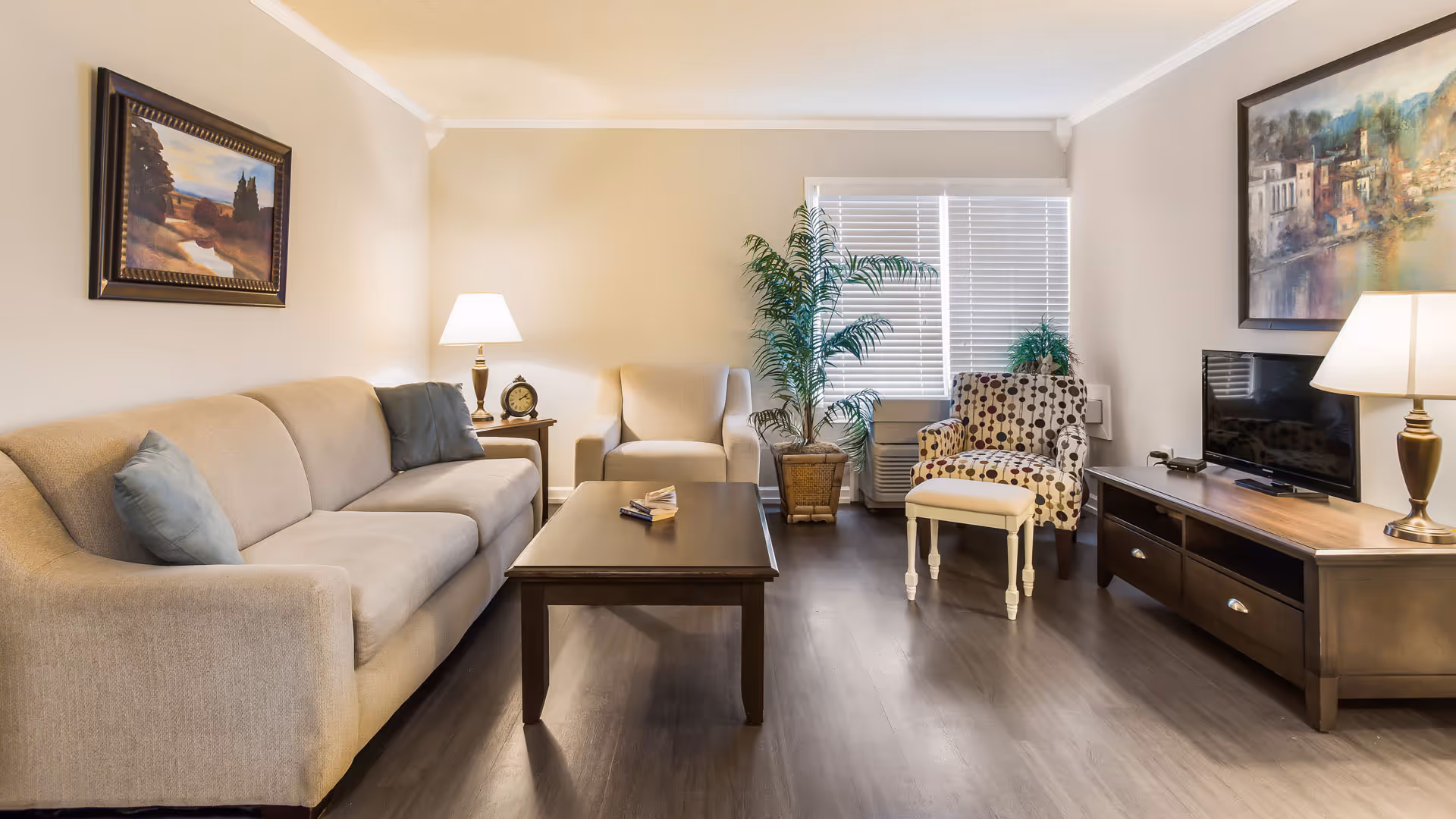 Bright furnished living room with a sofa, armchairs, coffee table, TV console, lamps, and potted plants.