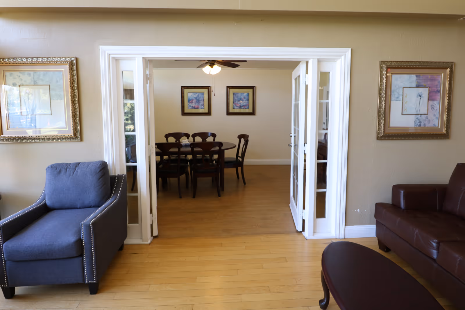 A living room with an armchair and sofa opening through French doors to a dining area with a table and chairs.