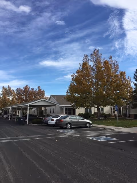 Parking lot with several cars parked under a covered area next to a single-story building surrounded by trees with autumn foliage under a partly cloudy sky.