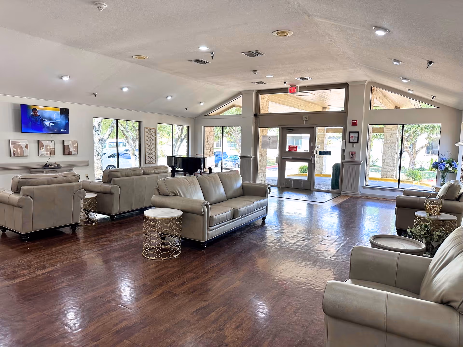 Spacious senior living lobby with leather sofas, side tables, a piano, a wall-mounted TV, and glass entrance doors.