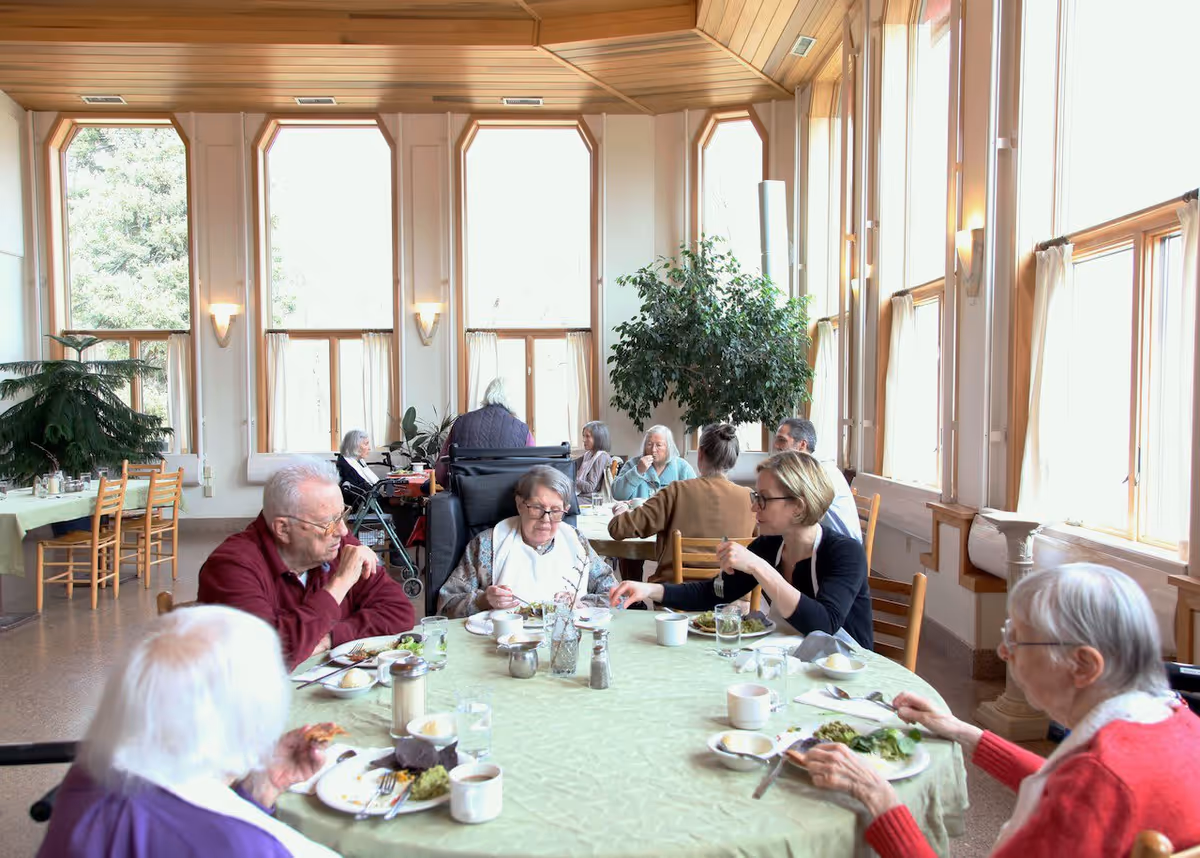 A group of elderly people and a caregiver sitting around a round table with a green tablecloth, eating a meal together in a bright dining room with large windows and tall plants.