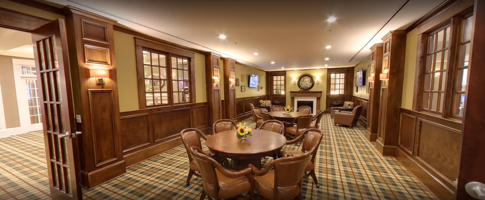 A cozy common area with round wooden tables and leather chairs arranged on a plaid carpet. The room features wood-paneled walls, wall sconces, two flat-screen TVs mounted on the walls, and a fireplace with a round mirror above it. The space is warmly lit with recessed ceiling lights.