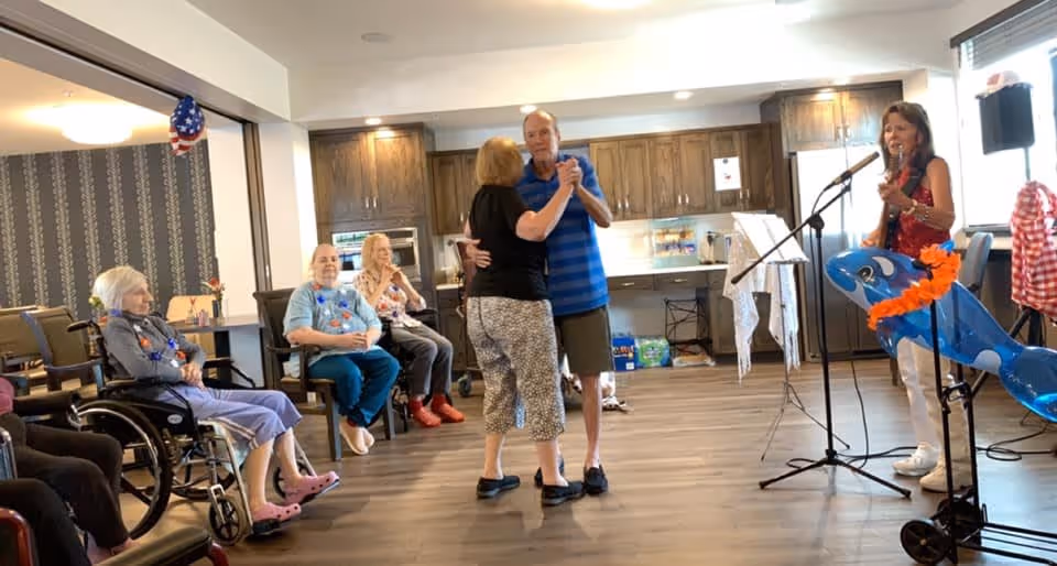 A group of elderly people in a common room at a senior living facility. Two people are dancing in the center while others are seated and watching. A woman is playing a guitar and singing into a microphone on the right side of the image. The room has wooden cabinets and a refrigerator in the background.