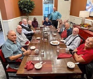 A group of elderly men sitting around a long wooden dining table in a dining room, with bowls and cups in front of them. One man is standing at the far end of the table, smiling. The room has warm-colored walls, a plant in the corner, and a red sideboard with decorative items.
