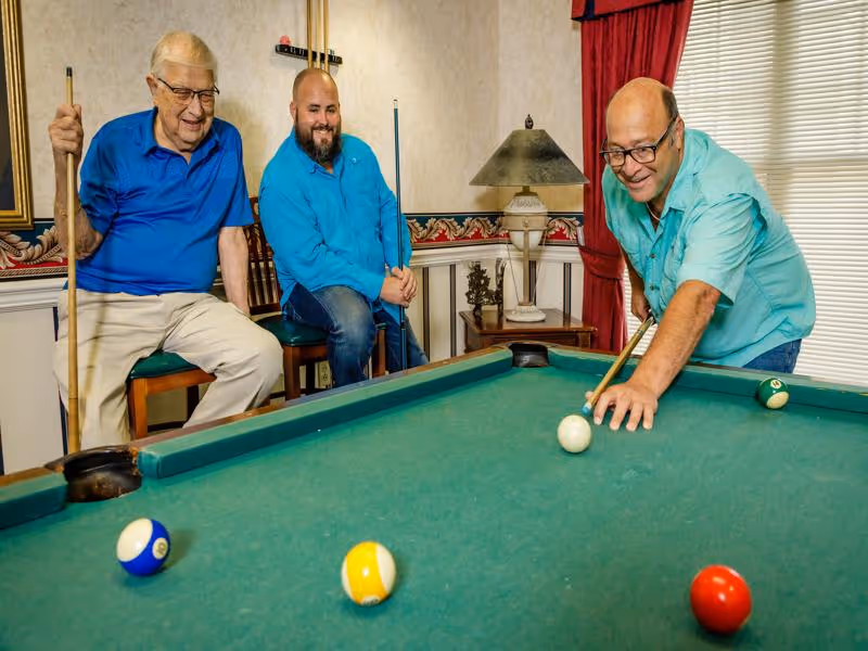 Three men playing pool in a room with a pool table. Two men are sitting on chairs holding pool cues, while the third man is leaning over the table aiming to hit the cue ball. The room has patterned wallpaper, a table lamp, and a window with blinds and red curtains.