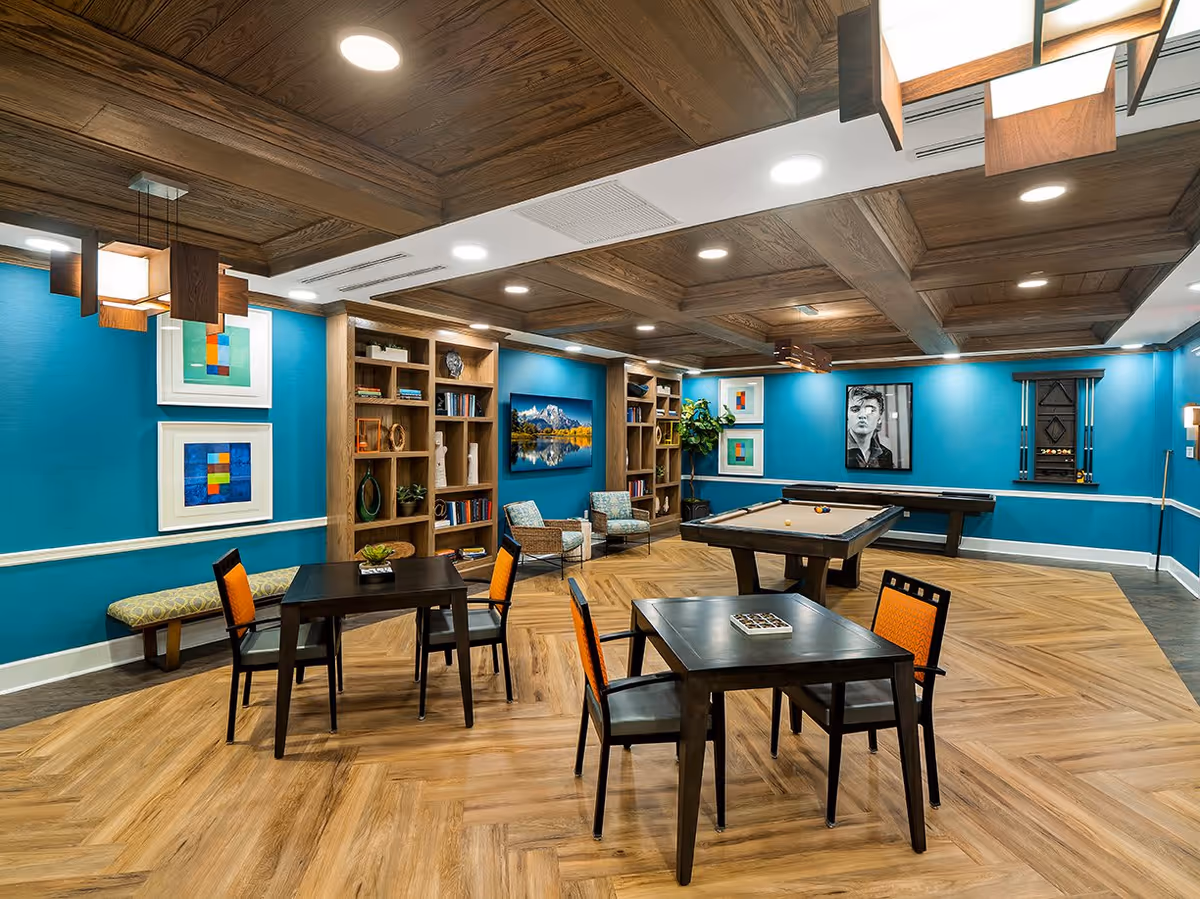 A stylish recreational room with blue walls and wooden ceiling beams. The room features two pool tables, two small square tables with chairs that have orange cushions, a bench with patterned upholstery, two armchairs, built-in wooden bookshelves with decorative items and books, framed abstract art and a black and white portrait on the walls, and a flat-screen TV displaying a mountain landscape.