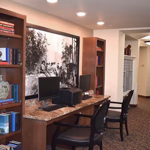 Interior view of a senior living facility's computer workstation area with two desktop computers on a marble countertop. Two black chairs are placed in front of the computers. Wooden bookshelves filled with books and decorative items flank the workstation. A large black and white photograph of people sitting outdoors is mounted on the wall above the countertop. The area is well-lit with recessed ceiling lights and has carpeted flooring.