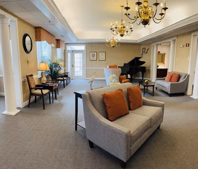 A spacious and well-lit common area with beige carpet and striped wallpaper. The room features two gray sofas with orange cushions, a black grand piano in the corner, a small table with a flower vase, several chairs along the wall, a clock, and two chandeliers hanging from the ceiling. There is a door with glass panels at the far end and some framed pictures on the wall.