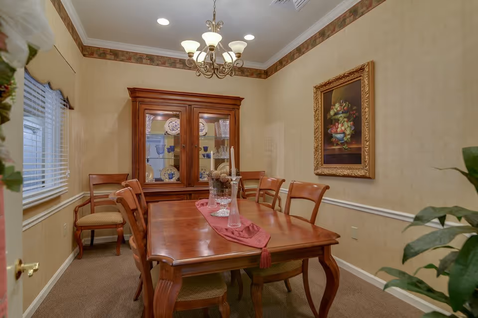 A traditional dining room with a wooden dining table and six matching chairs. The table has a red runner and decorative glass vases. A wooden china cabinet with glass doors displays plates and glassware. The room has beige walls with a decorative border near the ceiling, a chandelier hanging above the table, a window with blinds on the left, and a framed painting of fruit on the right wall.