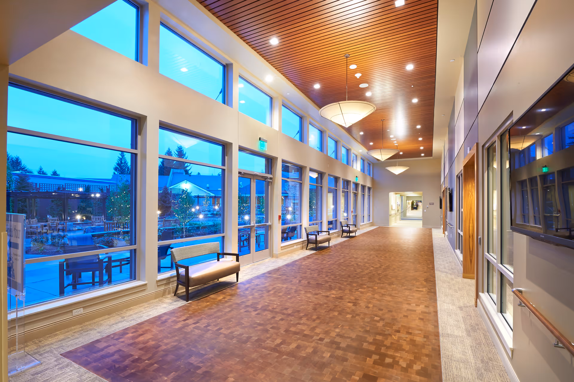 Spacious well-lit interior hallway with large windows, bench seating, a wood-paneled ceiling and pendant lights overlooking an outdoor courtyard.