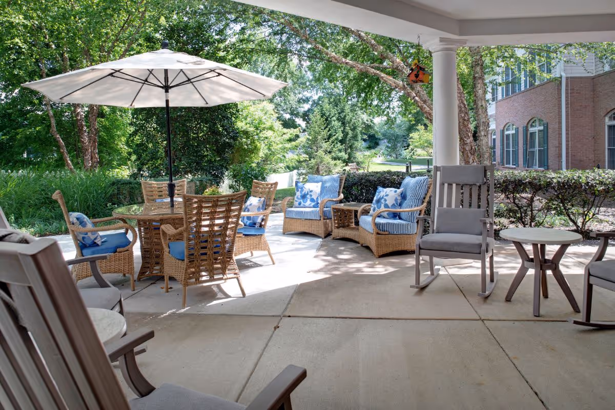 Covered outdoor patio with wicker and rocking chairs, a table with an umbrella, and cushioned seating overlooking landscaped greenery.