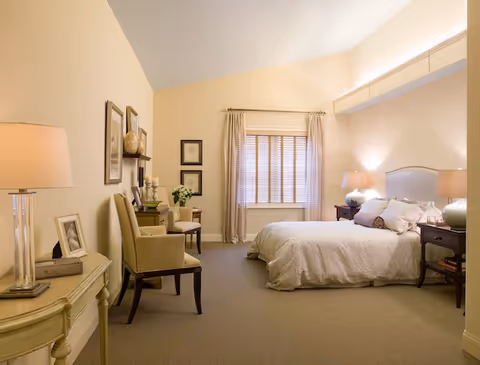 A cozy bedroom with a large bed covered in light-colored bedding, two bedside tables with lamps, a window with blinds and curtains, two upholstered chairs, framed artwork on the walls, and a small table with a lamp and picture frame.