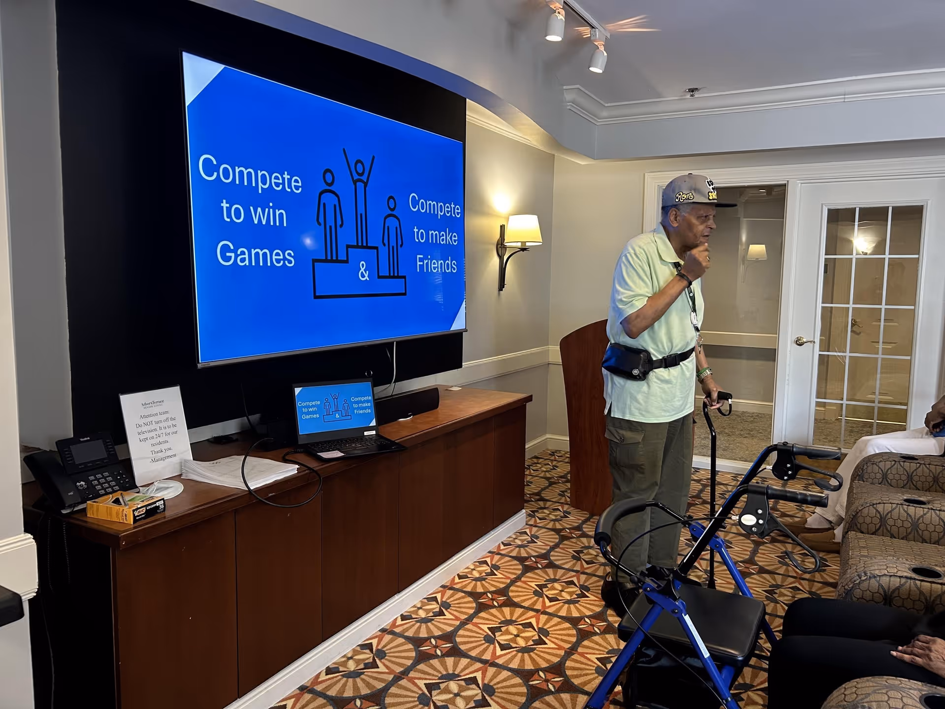 Elderly man with a walker stands in a seating area next to a large TV screen displaying a 'Compete to win Games' slide.