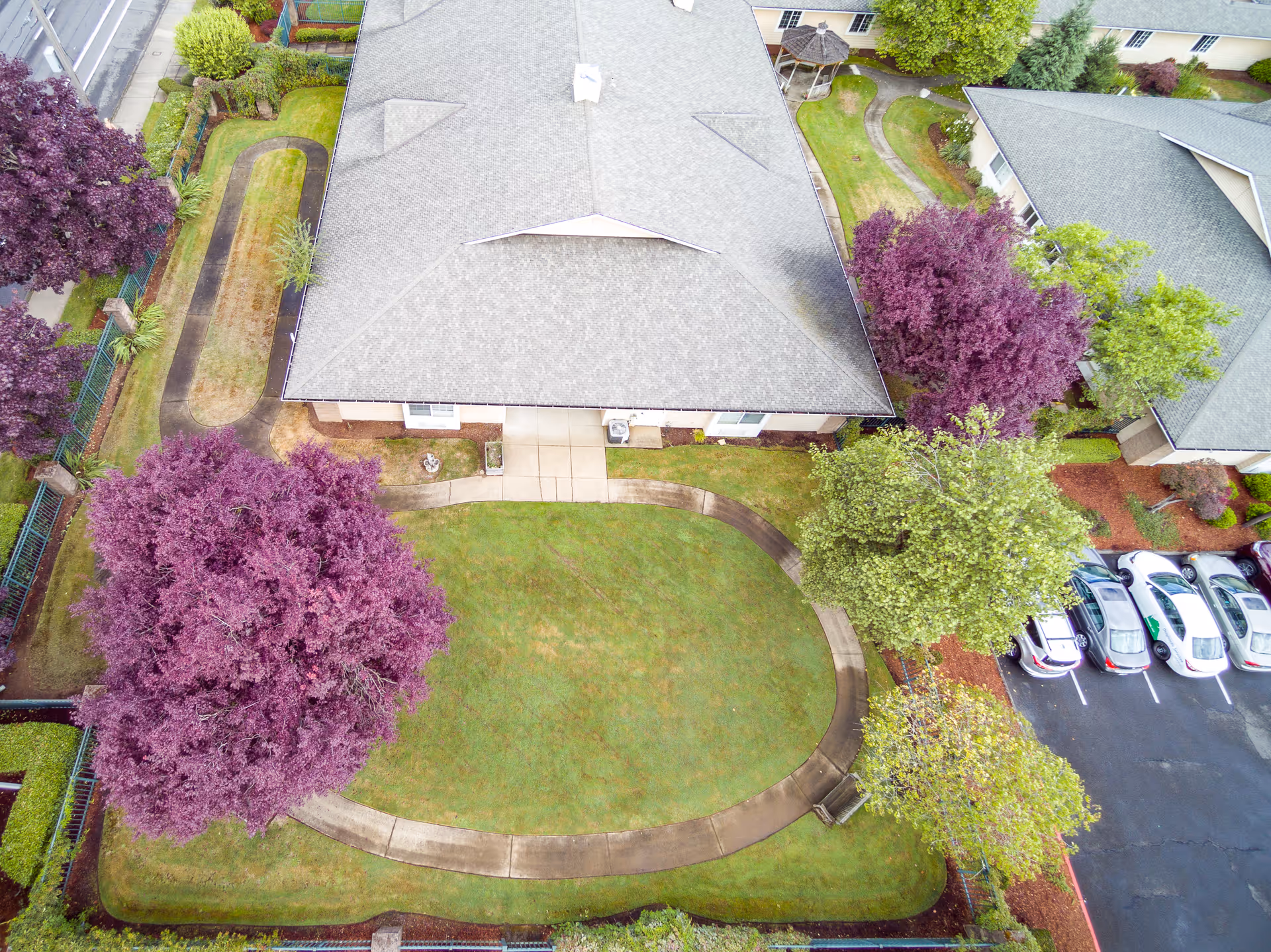 Aerial view of a senior living facility building with a gray roof surrounded by green lawns, purple and green trees, a curved concrete walkway, a small gazebo, and a parking lot with several cars.