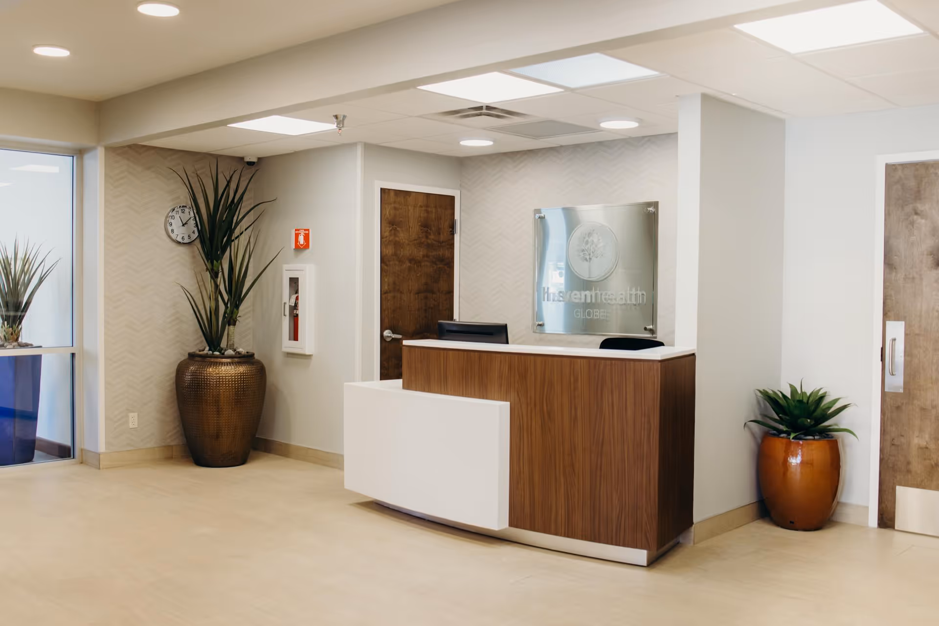 Reception area of Haven Health Globe facility featuring a wooden and white reception desk, two potted plants on either side, a wall clock, a fire extinguisher cabinet, and a sign with the facility's name on the wall behind the desk.