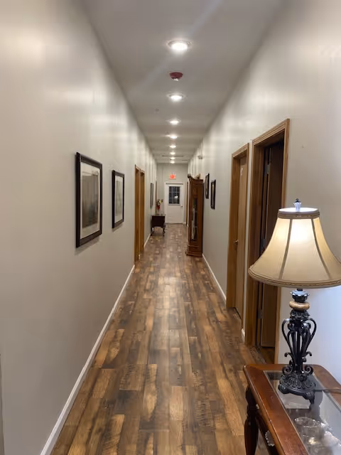 A long, narrow hallway with wooden flooring and white walls. Several framed pictures hang on the left wall, and multiple doorways are visible on the right. A decorative table with a lamp is positioned near the foreground on the right side. The hallway is well-lit with ceiling lights and leads to a door at the far end.