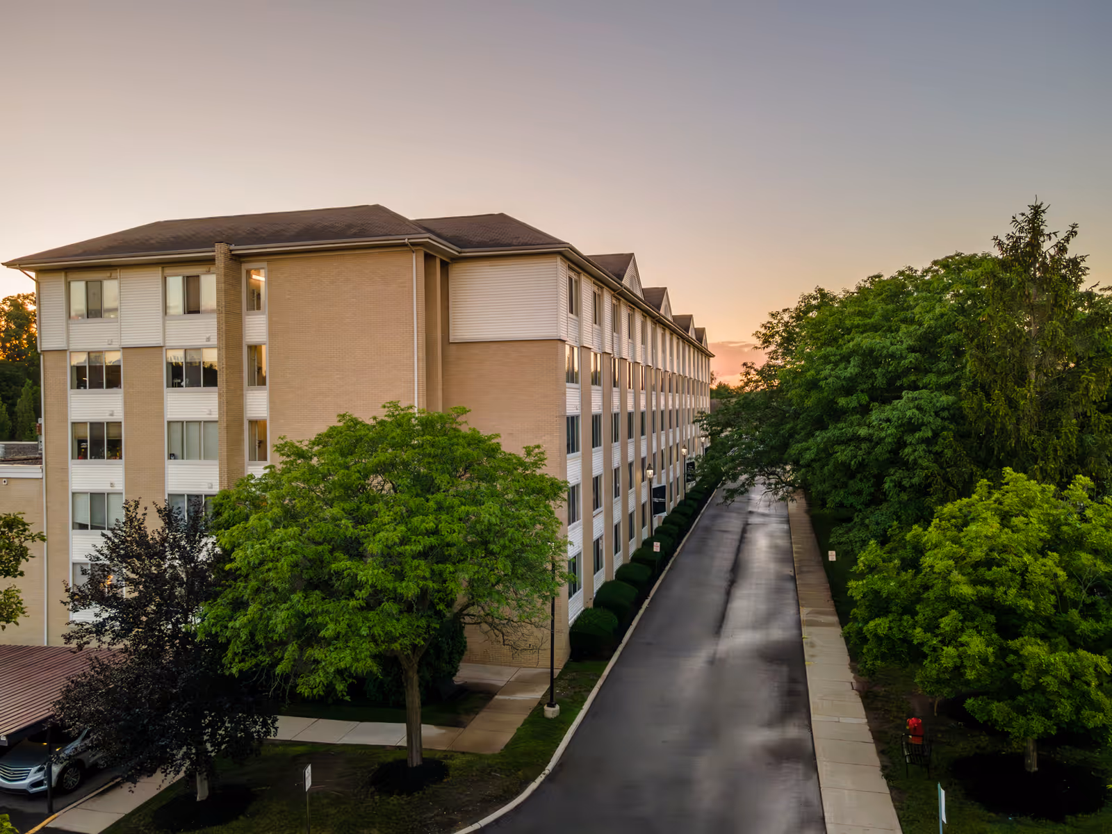 Exterior view of a multi-story senior living facility building with beige brick and white siding, surrounded by green trees and a paved driveway at sunset.