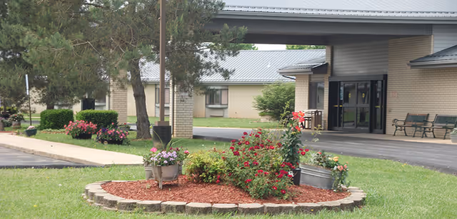 Exterior view of Lawrence County Residential Care Center showing the entrance with a covered drop-off area, benches near the entrance, a landscaped flower bed with various flowers and shrubs, and a paved driveway surrounded by grass and trees.