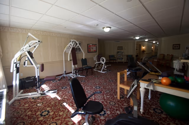 Small exercise room with weight machines, chairs, exercise balls, and other fitness equipment on a patterned carpet.