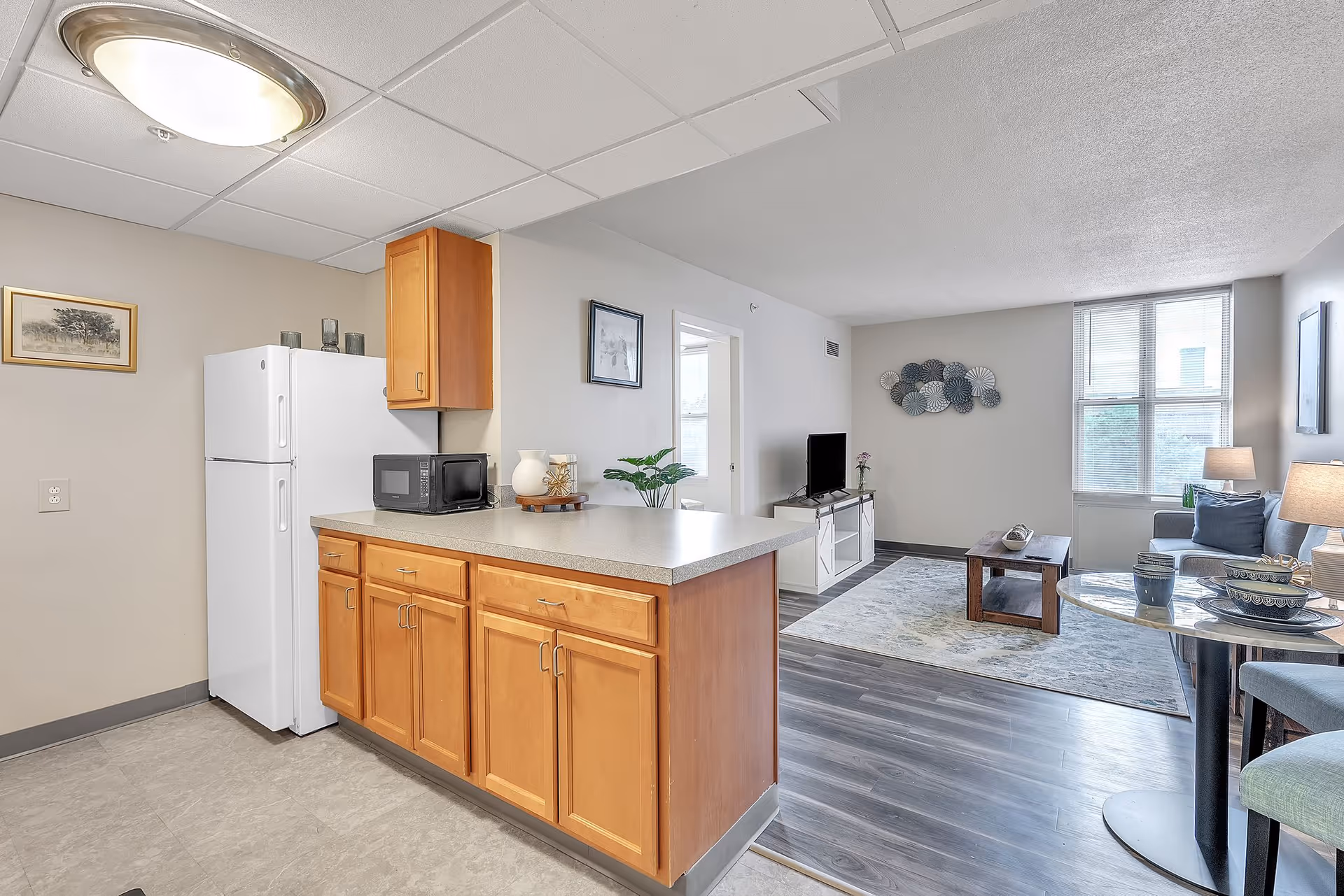 A bright and clean interior space showing a kitchen area with wooden cabinets, a white refrigerator, and a microwave on the counter. The kitchen opens into a living room with a TV on a white stand, a coffee table, a rug, a sofa with cushions, and a window letting in natural light. A small dining table with chairs and tableware is visible on the right side.