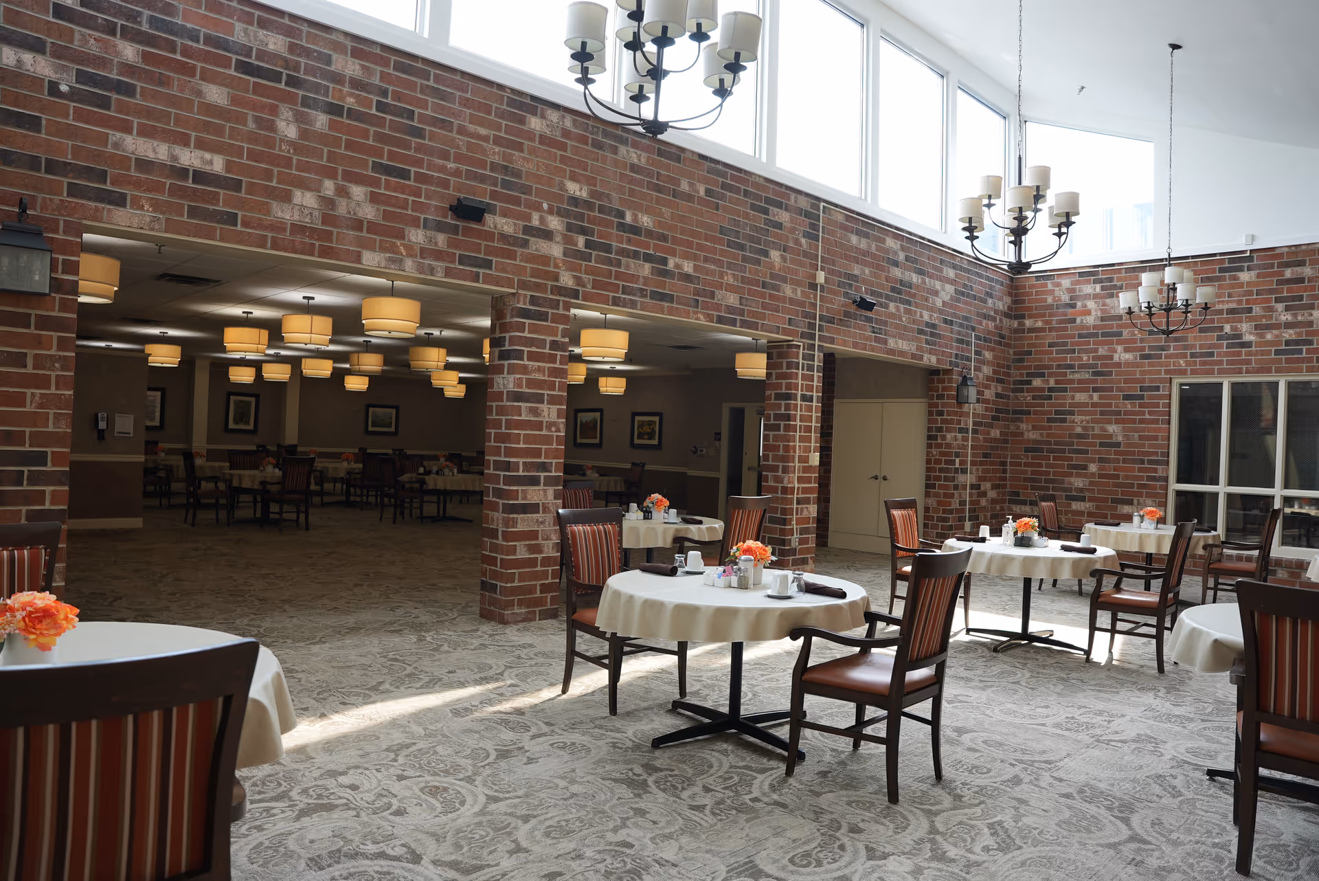 A bright dining area with round tables covered in white tablecloths, each set with cups, napkins, and small flower arrangements. The room features brick walls, large windows near the ceiling allowing natural light, and multiple chandeliers hanging from the ceiling. Beyond the dining area, there is an adjoining room with more tables and chairs under warm ceiling lights.