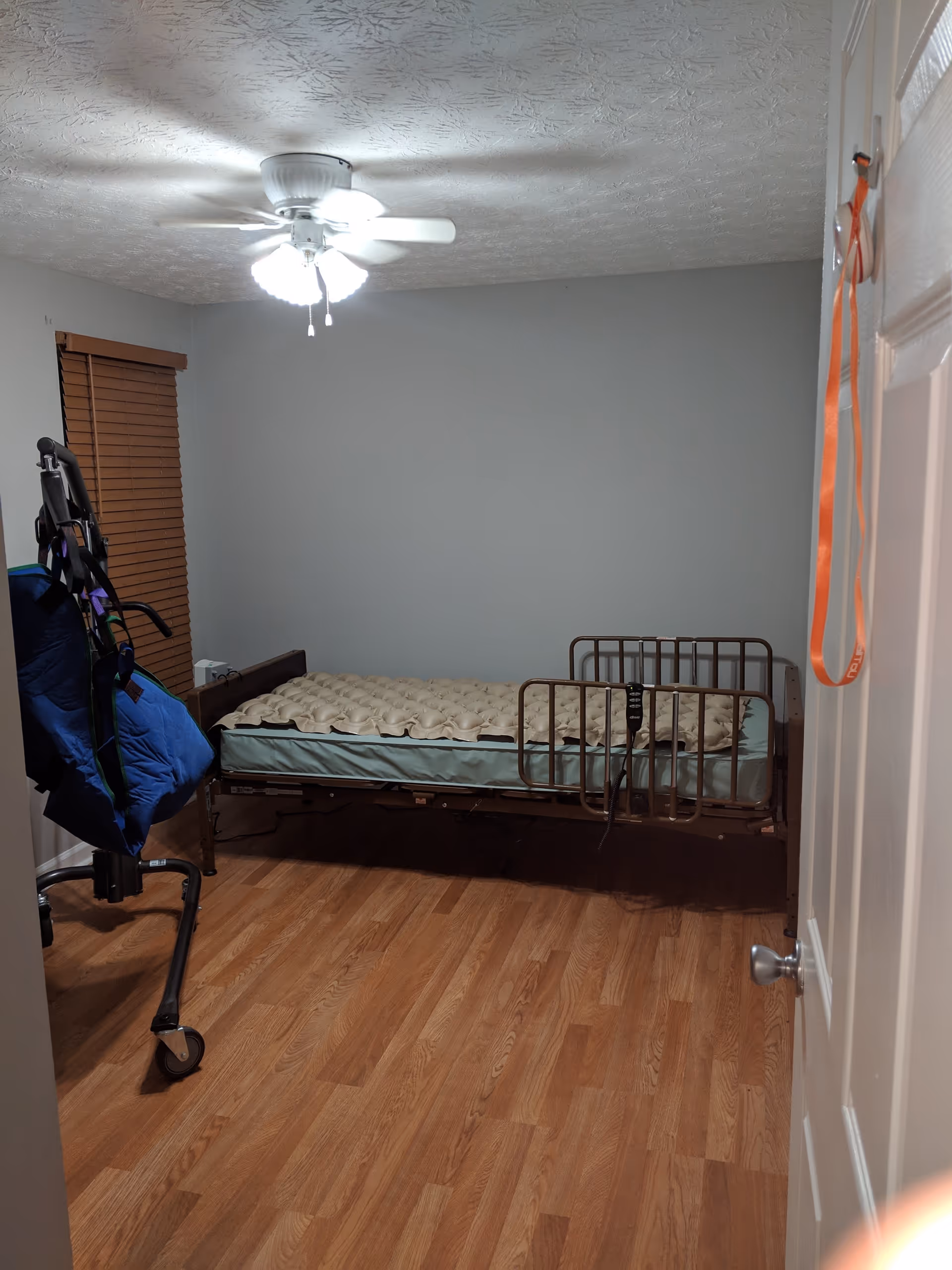 A simple bedroom with a medical bed featuring side rails and an air mattress. The room has wooden flooring, a ceiling fan with lights, a window with wooden blinds, and a blue patient lift device near the bed. An orange lanyard hangs on the white door.
