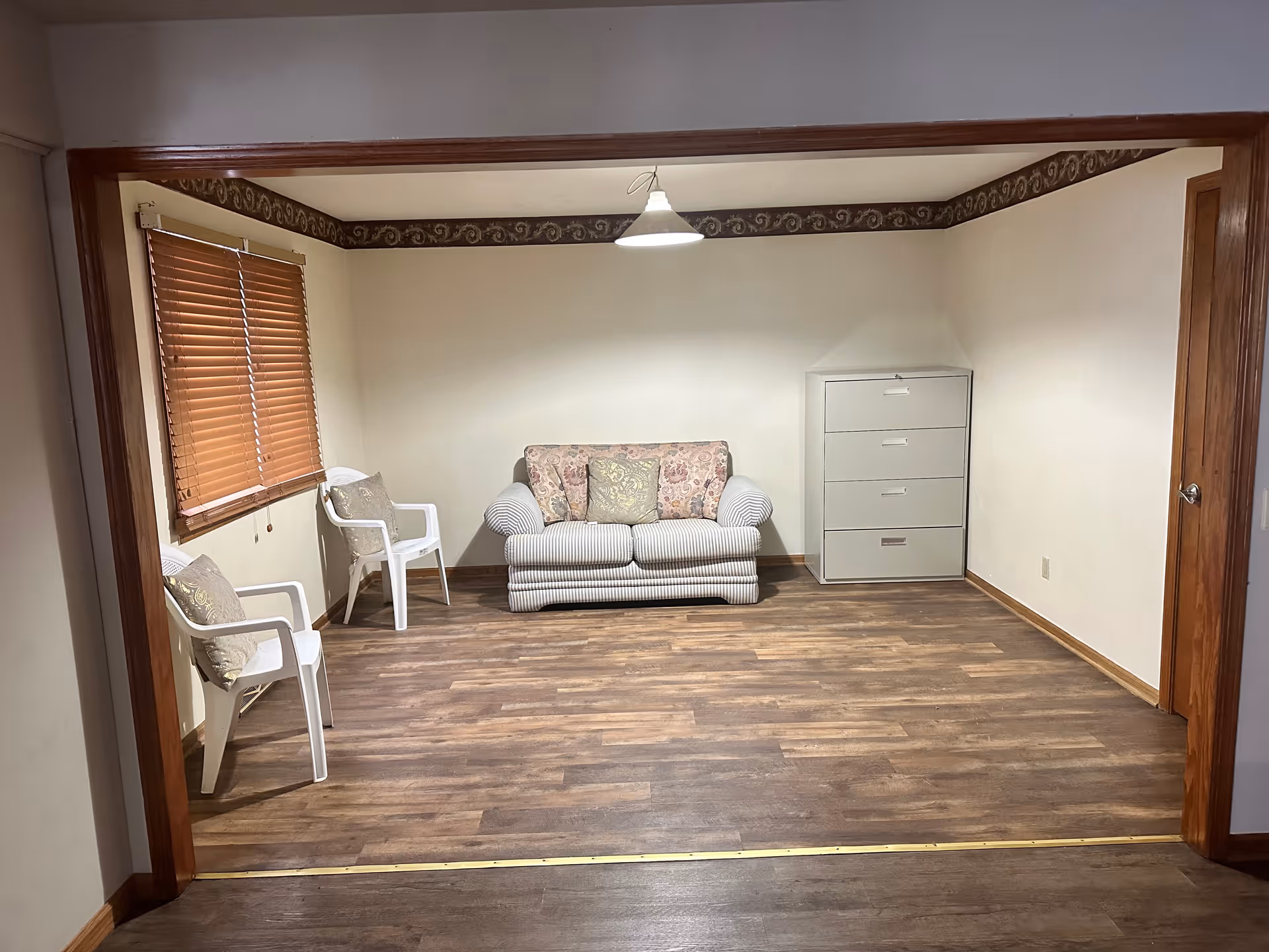 Small living room with a loveseat centered against the back wall, two white chairs by a window, and a gray filing cabinet to the right.