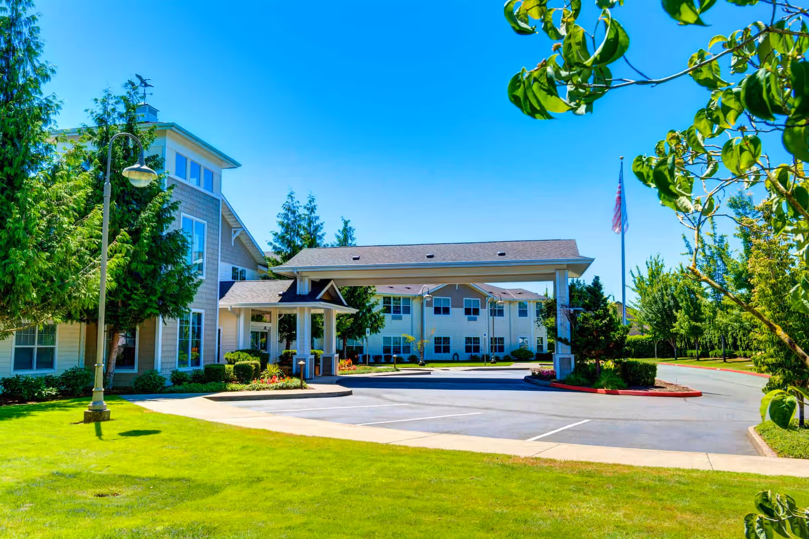 Exterior view of Clearwater Springs by Cogir facility showing a two-story building with a covered entrance, surrounded by green lawns, trees, and a clear blue sky.