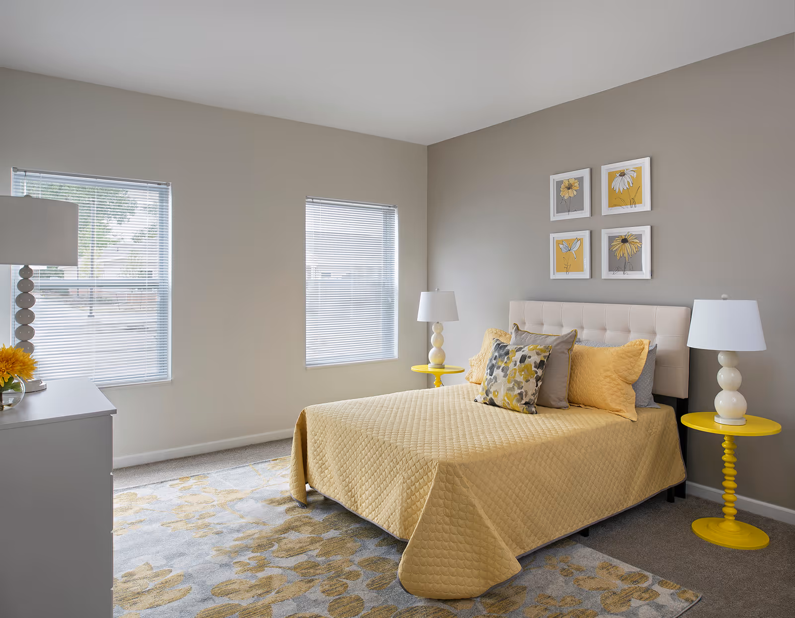 A bright bedroom with a bed covered in a yellow quilt and multiple pillows. There are two yellow side tables with white lamps on each side of the bed. The walls are painted light gray and decorated with four framed floral artworks. Two windows with blinds allow natural light into the room. A white dresser with a lamp and a vase of yellow flowers is visible on the left side.