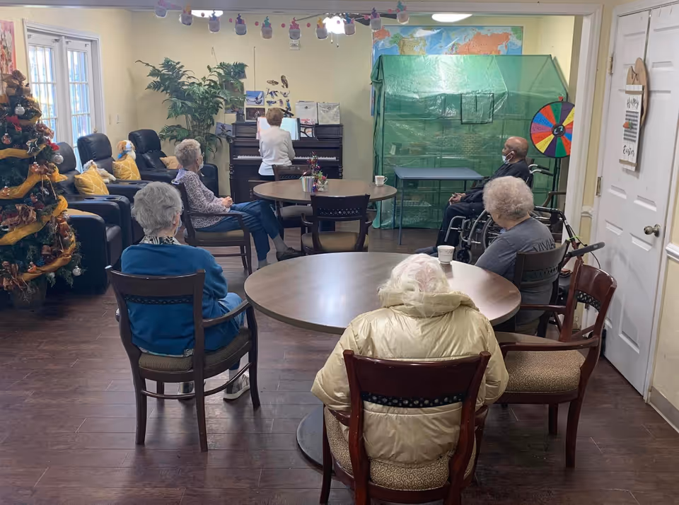 A group of elderly people seated around tables in a common room, facing a woman playing a piano. The room has a Christmas tree decorated with ornaments and ribbons, a large plant, and a colorful wheel on a stand near the back wall. The floor is wooden, and the walls are light-colored with some decorations.