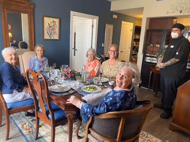 Five elderly women sit around a set dining table enjoying a meal while a masked staff member stands nearby in a dining room.