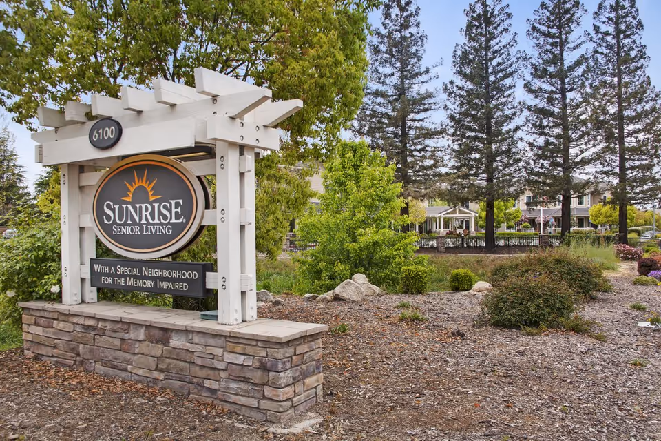 Outdoor view of the Sunrise Senior Living facility sign with landscaping, trees, and the building visible in the background.