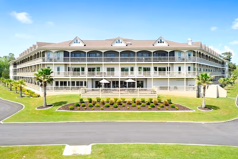 Exterior view of a large three-story senior living facility building with multiple balconies, surrounded by a well-maintained lawn, palm trees, and a paved driveway under a blue sky with some clouds.