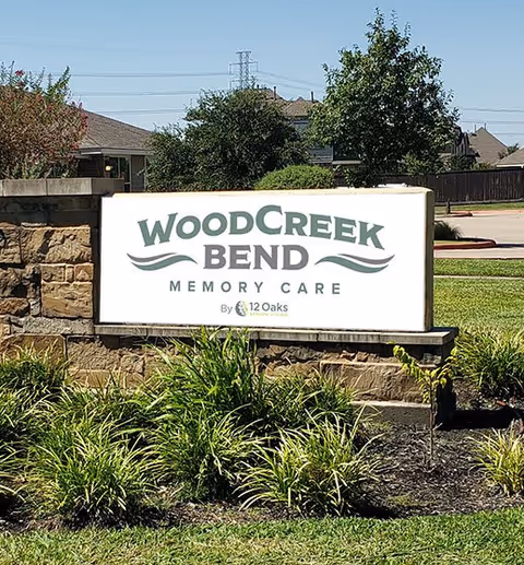 Outdoor sign for WoodCreek Bend Memory Care by 12 Oaks, surrounded by landscaping with green plants and grass, with a stone base and a clear sky in the background.